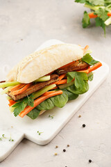Traditional vietnamese banh mi sandwich with sliced smoked tofu, fresh spinach, shredded carrots and peeled cucumbers, cilantro on marble board on light grey textured background