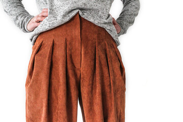 A white woman stands in front with her hands on her waist, dressed in a jumper and terracotta trousers. Details of everyday clothes. On a white background. Part of the body is visible in the waist