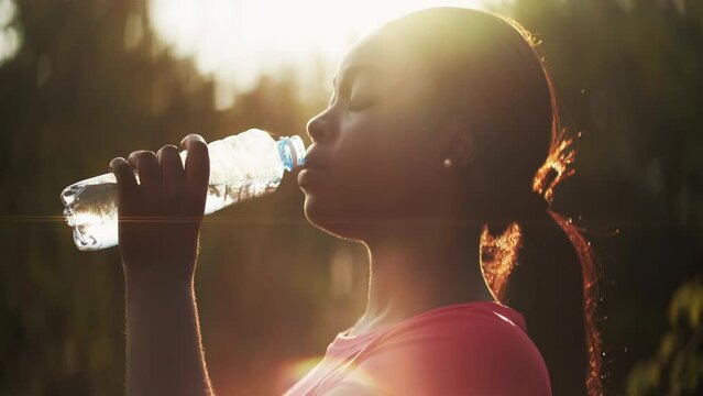 Thirsty woman. Body refreshment. Healthy hydration. Fitness break. Happy athletic girl drinking water in defocused park lens flare landscape.
