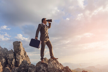 Businessman using business attache and binoculars standing on top of cliff in summer mountains enjoying Sunset