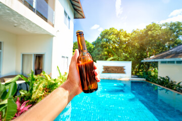 Vacation concept. Male hand holding bottle of beer against swimming pool.