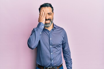 Middle age man with beard and grey hair wearing business clothes covering one eye with hand, confident smile on face and surprise emotion.