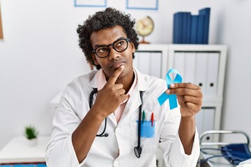 African doctor man holding blue ribbon serious face thinking about question with hand on chin, thoughtful about confusing idea