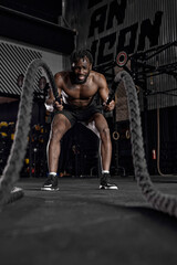 Fototapeta na wymiar fit active afro man with battle rope doing exercise in functional training fitness gym exercising with battle ropes at gym. black shirtless guy working out arms and cardio for cross fit