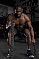 Fototapeta na wymiar Young black man doing strength training using battle ropes at modern dark gym. Athlete moving the ropes in wave motion as part of fat burning workout, active intense cross fit training, exercising