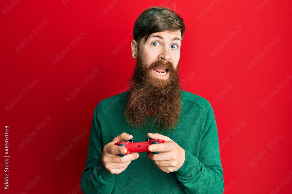 Sticker Redhead man with long beard playing video game holding controller clueless and confused expression. doubt concept.