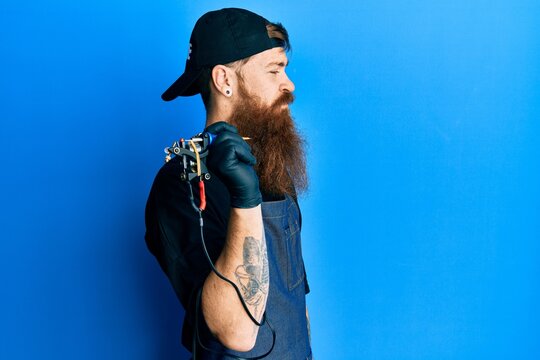 Redhead man with long beard tattoo artist wearing professional uniform and gloves looking to side, relax profile pose with natural face and confident smile.