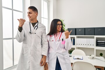 Obraz premium Young doctors wearing uniform and stethoscope at the clinic pointing thumb up to the side smiling happy with open mouth