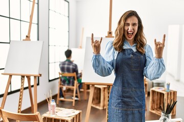 Young artist woman at art studio shouting with crazy expression doing rock symbol with hands up....