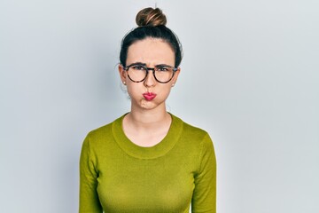 Young hispanic girl wearing casual clothes and glasses puffing cheeks with funny face. mouth inflated with air, crazy expression.