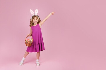Obraz na płótnie Canvas Full body little beautiful smiling girl in Easter bunny ears holds a basket with eggs on pink background and pointing away with hand. Copy space for text
