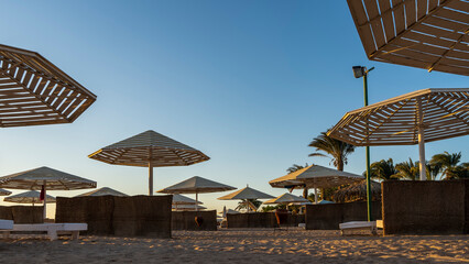 Morning on the Red Sea beach. Lattice umbrellas from the sun and palm trees against the blue sky. There are empty deck chairs and wicker fences on the sand. Egypt