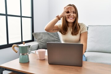 Young brunette woman using laptop at home drinking a cup of coffee doing ok gesture with hand smiling, eye looking through fingers with happy face.