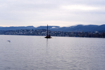 Fototapeta na wymiar Beautiful scenic landscape with Lake Zurich and working raft in the foreground and Swiss Alps in the background on a cloudy winter afternoon. Photo taken February 3rd, 2022, Zurich, Switzerland.