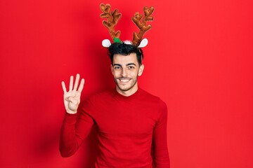Young hispanic man wearing cute christmas reindeer horns showing and pointing up with fingers number four while smiling confident and happy.