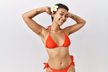Young hispanic woman with short hair wearing bikini relaxing and stretching, arms and hands behind...