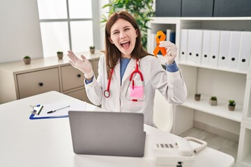 Young doctor woman holding awareness orange ribbon celebrating achievement with happy smile and...