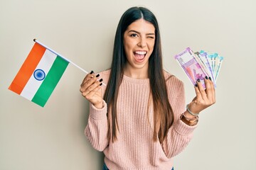 Young hispanic woman holding india flag and rupee banknotes winking looking at the camera with sexy...