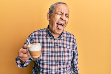 Handsome senior man with grey hair drinking a cup coffee sticking tongue out happy with funny expression. emotion concept.