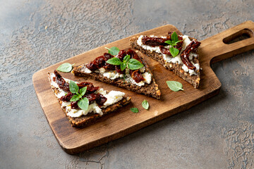 Bruschetta or whole grain bread sandwich with cream cheese and sun-dried tomatoes. Crostini on a wooden serving board on a dark kitchen table closeup