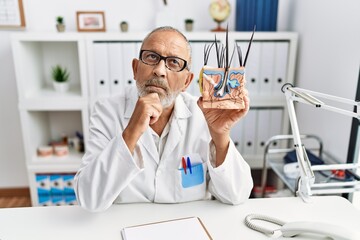 Mature doctor man holding model of human anatomical skin and hair serious face thinking about question with hand on chin, thoughtful about confusing idea