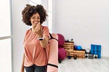 African american woman with afro hair holding yoga mat at pilates room beckoning come here gesture with hand inviting welcoming happy and smiling