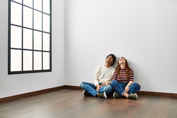 Young hispanic couple looking to the ceiling sitting on the floor at empty new home.