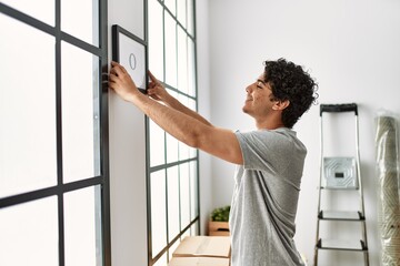 Young hispanic man smiling happy hanging frame at new home.