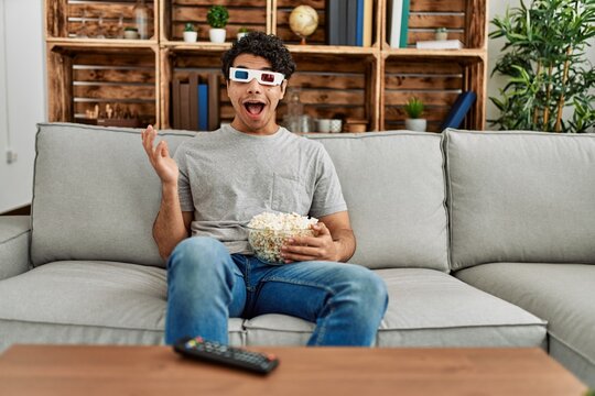 Young hispanic man watching movie using 3d glasses sitting on the sofa at home.