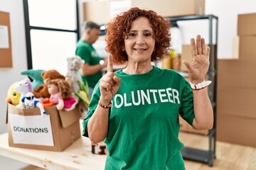 Middle age woman wearing volunteer t shirt at donations stand showing and pointing up with fingers...