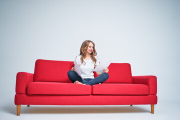 Work and relaxation. Pretty young woman reading documents while sitting on red sofa on white background.