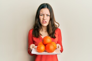 Young brunette woman holding plate with fresh oranges clueless and confused expression. doubt...