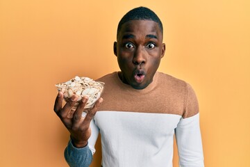 Young african american man holding bowl with pumpkin seeds scared and amazed with open mouth for...