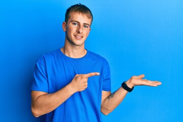 Young caucasian man wearing casual blue t shirt amazed and smiling to the camera while presenting with hand and pointing with finger.