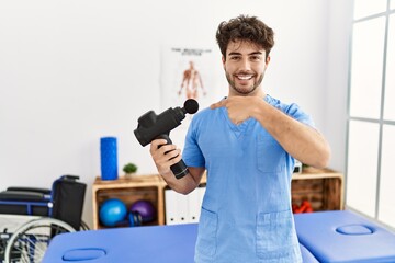 Hispanic physiotherapy man working at pain recovery clinic with muscle gun smiling happy pointing with hand and finger