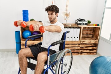 Young hispanic man sitting on wheelchair doing rehab using dumbbells at clinic