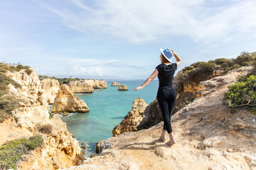 a young beautiful girl in a blue hat stands on a rock against the background of the beach and the ocean in Portugal