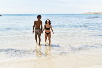 Young interracial tourist couple wearing swimwear walking with hands together at the beach.