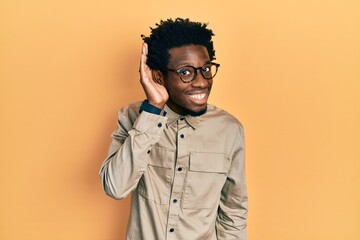 Young african american man wearing casual clothes and glasses smiling with hand over ear listening an hearing to rumor or gossip. deafness concept.