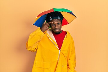 Young african american man wearing yellow raincoat pointing unhappy to pimple on forehead, ugly...