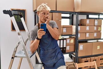 Young blond man using smartphone working at storehouse looking stressed and nervous with hands on...