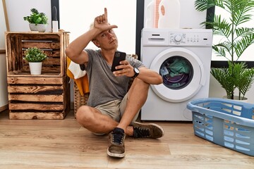 Young blond man doing laundry using smartphone making fun of people with fingers on forehead doing...