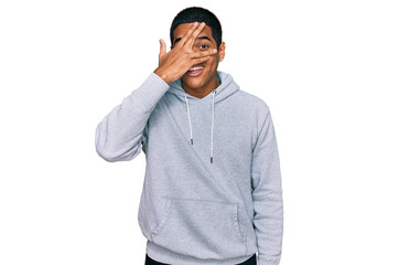 Young handsome hispanic man wearing casual sweatshirt peeking in shock covering face and eyes with hand, looking through fingers with embarrassed expression.