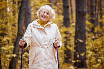 Senior aged woman standing with nordic walking poles in autumn park. Healthy lifestyle concept. Mature caucasian female resting after exercise outdoors, laughing smiling, enjoying time outdoors