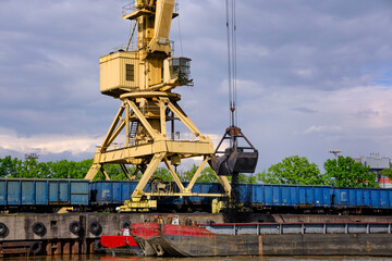 Fototapeta na wymiar River port crane with clamshell or griper loading coal to river drag boats or barges moored by pier on cloudy day. Energy, minerals, environment 