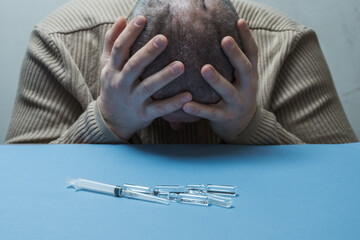 The man hides his face with his hands, syringes and ampoules are lying in front of him on the table.