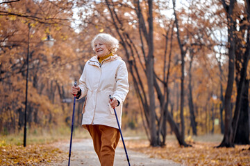 Fit happy gray haired mature female in coat enjoying health-promoting physical activity using walking poles having excited joyful facial expression, breathing fresh air in autumn ature, smiling