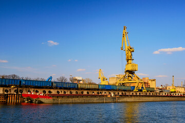 River port crane loading open-top gondola cars on sunny day. Empty river drag boats or barges moored by pier, Empty cars ready for loading.