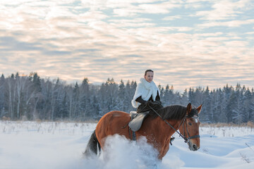 A girl in a white cloak rides a brown horse in winter.