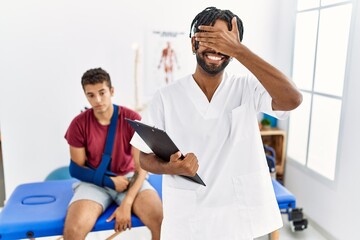 Obraz premium Young hispanic man working at pain recovery clinic with a man with broken arm smiling and laughing with hand on face covering eyes for surprise. blind concept.